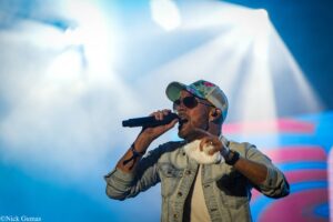 Male singer on stage, singing into a handheld microphone, wearing a floral cap, sunglasses, and a denim jacket with a bright blue-pink backdrop.