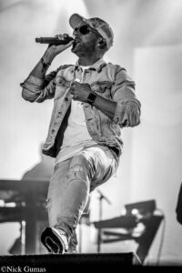 Black-and-white image of amale singer wearing a denim jacket and cap, singing into a handheld microphone on stage.
