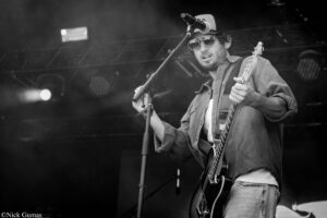 Black-and-white photo of a male guitarist on stage, singing into a mic while playing an electric guitar.