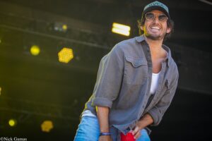 Smiling man on stage wearing a blue cap and sunglasses, holding a red cup, denim shirt visible in the foreground.