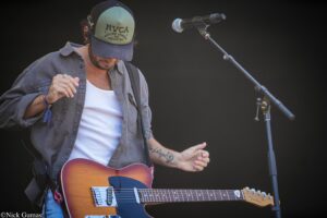 Musician playing an orange Fender Telecaster on stage, wearing a blue denim shirt and RVCA cap, microphone to his right.