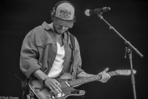 Male guitarist wearing a cap and denim shirt, playing an electric guitar on stage with a mic stand nearby (black-and-white photo).