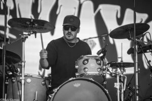 Drummer wearing sunglasses and a cap, gripping drumsticks behind a loud drum kit on stage (black-and-white photo).