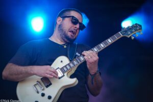 Guitarist wearing sunglasses and a black shirt plays a cream-colored electric guitar on stage under blue lights.