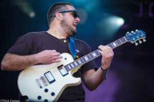 Guitarist performing on stage, wearing sunglasses and a gold necklace, playing a white electric guitar.