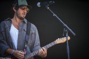 Male guitarist performing on stage, wearing a green cap and sunglasses, strumming an electric guitar with a microphone nearby