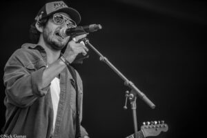 Male singer wearing glasses and a cap performs into a microphone on stage, a guitar visible in the background (black-and-white).