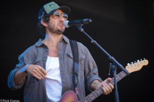 Male guitarist and singer on stage, wearing sunglasses and a cap, singing into a microphone while strumming an electric guitar.