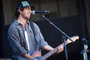 Male guitarist singing into a microphone during a live performance, wearing sunglasses and a gray shirt on stage.