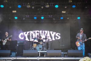 Rock band performing on an outdoor stage: guitarist left, drummer center, guitarist/singer right, blue stage lighting and a large stylized banner behind them.