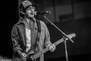 Male guitarist wearing a cap and sunglasses sings into a microphone on stage while playing an electric guitar in a live performance (black and white).