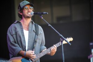 Male guitarist performing on stage, singing into a microphone while playing an electric guitar, wearing a cap, sunglasses and a gray shirt.