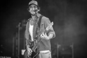 Male guitarist singer performing on stage with microphone, wearing a cap and sunglasses.