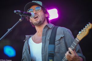 Male singer wearing sunglasses and a cap performs on stage into a microphone while playing an electric guitar under purple lights.