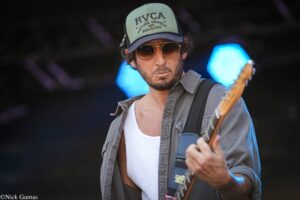 Male guitarist wearing a green RVCA cap and sunglasses, playing an electric guitar on stage.