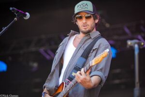 Male guitarist on stage wearing a green cap and sunglasses, playing an electric guitar in a live performance.