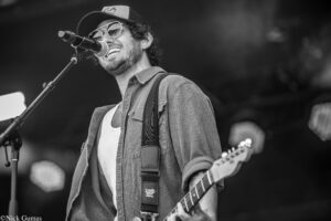 Musician wearing a cap and sunglasses sings into a microphone while playing an electric guitar on stage.