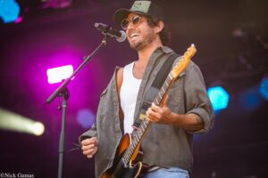 Male musician wearing a cap and sunglasses, singing into a microphone while playing an electric guitar on a colorful stage.1