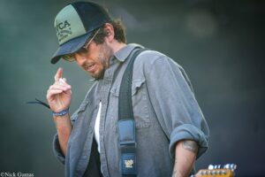 Male musician on stage wearing a green-and-black trucker cap, sunglasses, and a gray shirt, looking down as he holds his hand near his mouth.