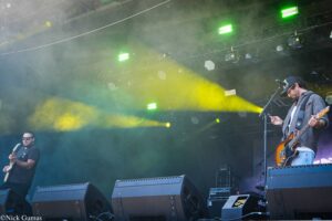 Two guitarists perform on a smoky outdoor stage with green and yellow lights and large speakers in front.