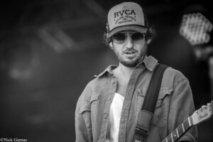 Black-and-white photo of a man wearing an RVCA cap and sunglasses, playing a guitar with a strap over his shoulder.
