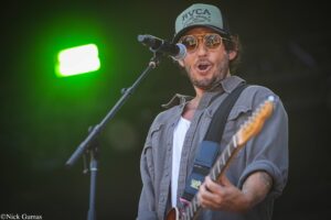 Male guitarist and vocalist on stage, wearing a gray shirt and RVCA cap, singing into a microphone with mirrored sunglasses reflecting the crowd; bright green stage light in background.
