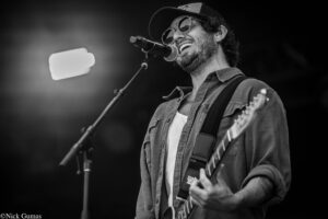 Male musician wearing sunglasses and a cap, singing into a microphone while playing an electric guitar on stage (black-and-white).
