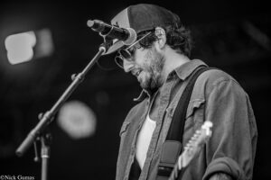 Male musician with a beard wearing a cap and sunglasses, singing into a microphone while playing an electric guitar on stage.