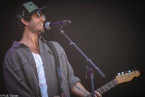 Male musician singing into a microphone while playing an electric guitar on stage, wearing a cap and sunglasses.