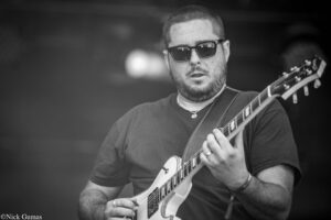 Male guitarist wearing sunglasses, playing an electric guitar on stage.