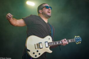 Guitarist wearing sunglasses and a black shirt plays a white electric guitar on stage, mouth open in performance.