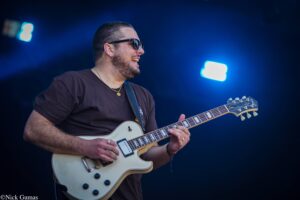 Male guitarist wearing sunglasses plays a cream-colored electric guitar on a dark stage with blue lighting behind him.