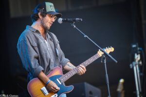 Male guitarist performing on stage, wearing hat and sunglasses, singing into a mic while playing a sunburst electric guitar.