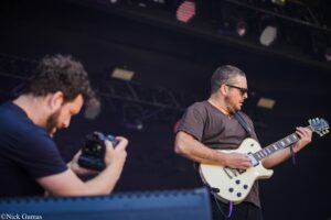 Guitarist in a brown shirt and sunglasses playing a white electric guitar on stage; a photographer is visible in the foreground photographing him.