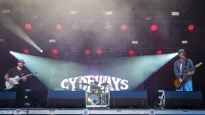 Two guitarists performing on an outdoor stage with a drummer in back, red stage lights and a large banner behind them.