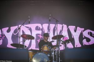 Drummer on stage behind a tan drum kit with two overhead mics, pink graffiti backdrop visible behind.