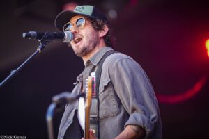 Male musician singing into a microphone on stage, wearing a cap, sunglasses, and a guitar strapped on.