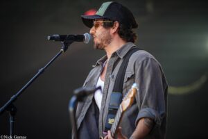 Male musician wearing a cap and sunglasses singing into a microphone while playing an electric guitar on stage, with a dark blurred background.