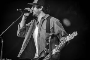 Male singer performs on stage, gripping a microphone while playing an electric guitar, wearing a cap and glasses in a black-and-white photo.