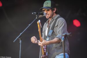 Male musician wearing a green and black cap and sunglasses, singing into a microphone while playing an electric guitar on stage