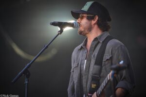 Male musician with a beard and cap sings into a microphone while playing a guitar on stage, spotlight behind him