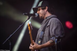 Male guitarist and vocalist performing on stage, singing into a microphone with a cap and sunglasses on.