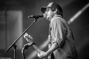 Male singer wearing a cap and glasses, strumming a guitar and singing into a microphone on stage (black and white).