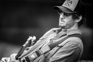 Man wearing a cap and sunglasses, playing an electric guitar in a black-and-white photo.