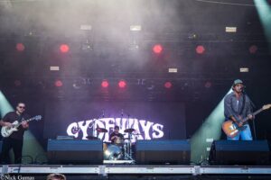 Three-member rock band performs on an outdoor stage with red spotlights and fog, guitarist left, bassist/singer right, drummer at center behind drums behind a large banner.