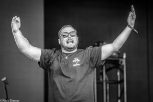 Male performer on stage with arms raised, wearing sunglasses and a Goleta t-shirt.