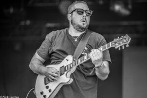 Male guitarist wearing sunglasses, playing a white electric guitar on stage.