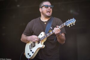 Man wearing dark sunglasses and a brown t-shirt plays a cream-colored electric guitar on stage, mouth open as if singing or shouting, with a black backdrop behind.