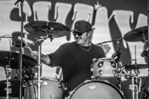 Drummer wearing a cap and sunglasses performs behind a drum set on stage, mid-performance.
