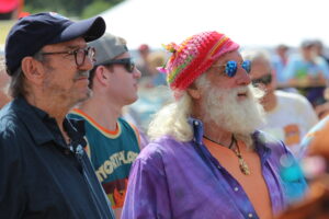 Three men at an outdoor festival: foreground left wearing a dark shirt and cap, middle man in teal shirt, and a bearded man on the right with a bright knitted hat and blue mirrored sunglasses.